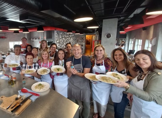 Alunos e o Chef Giuseppe Gerundino posam juntos segurando pratos de massa recém-preparados durante uma aula prática na cozinha.