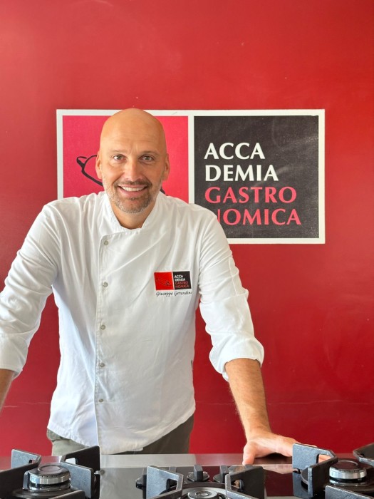 Chef Giuseppe Gerundino, fundador da Accademia Gastronomica, sorrindo e vestido com sua dólmã branca em frente ao logo da escola.